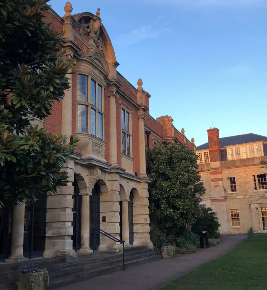 Widok na zewnątrz biblioteki Somerville College z czerwoną, ceglaną elewacją w stylu wiktoriańskim, kamiennymi łukowymi wejściami, ozdobnymi szczytami, dużymi wielodzielnymi oknami oraz magnoliami rosnącymi wzdłuż ścieżki na trawniku.