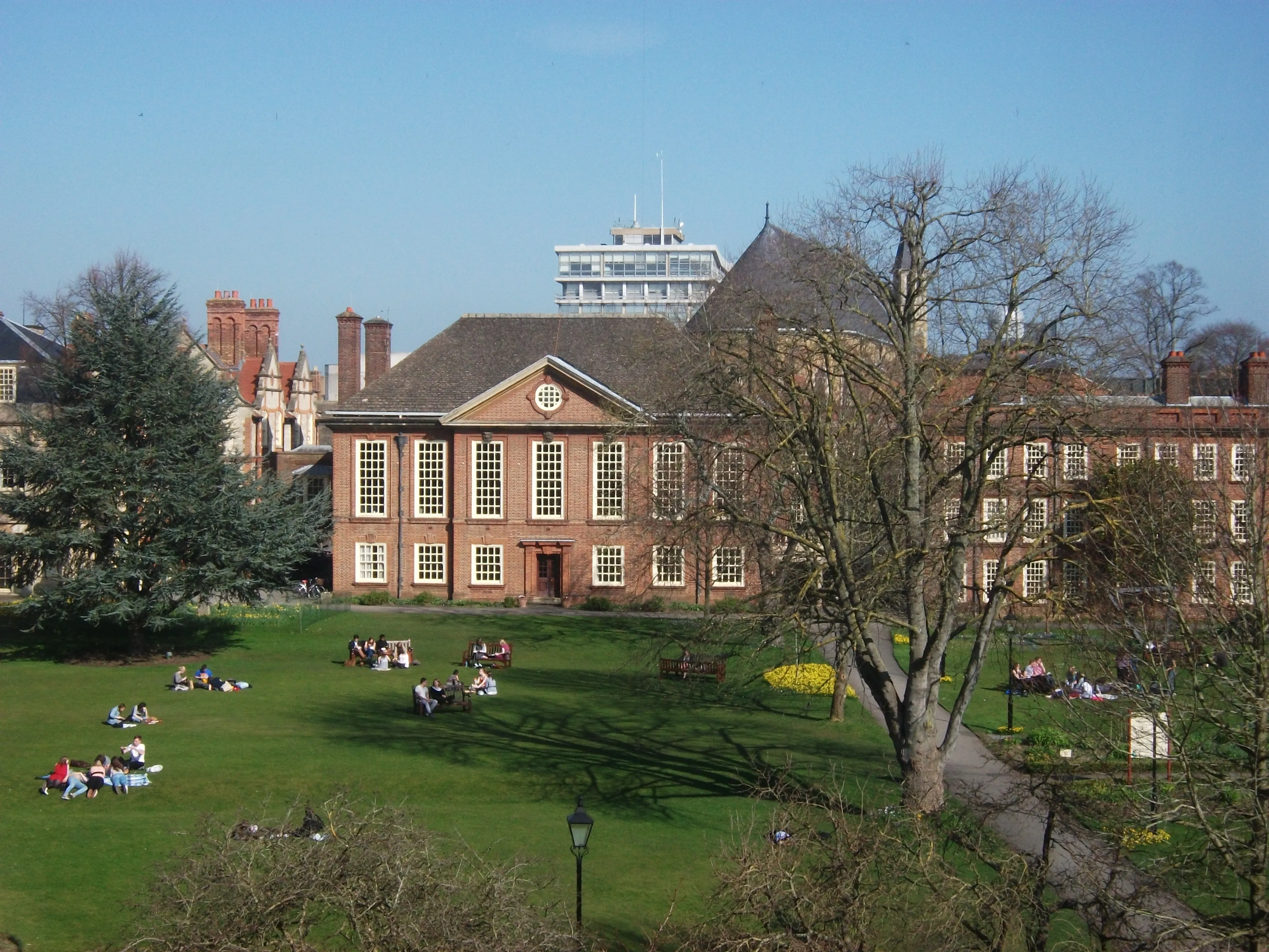 Główny dziedziniec Somerville College przedstawiający budynek z czerwonej cegły z dużymi oknami skrzynkowymi, zielony trawnik z siedzącymi ludźmi oraz drzewa pod bezchmurnym niebieskim niebem.