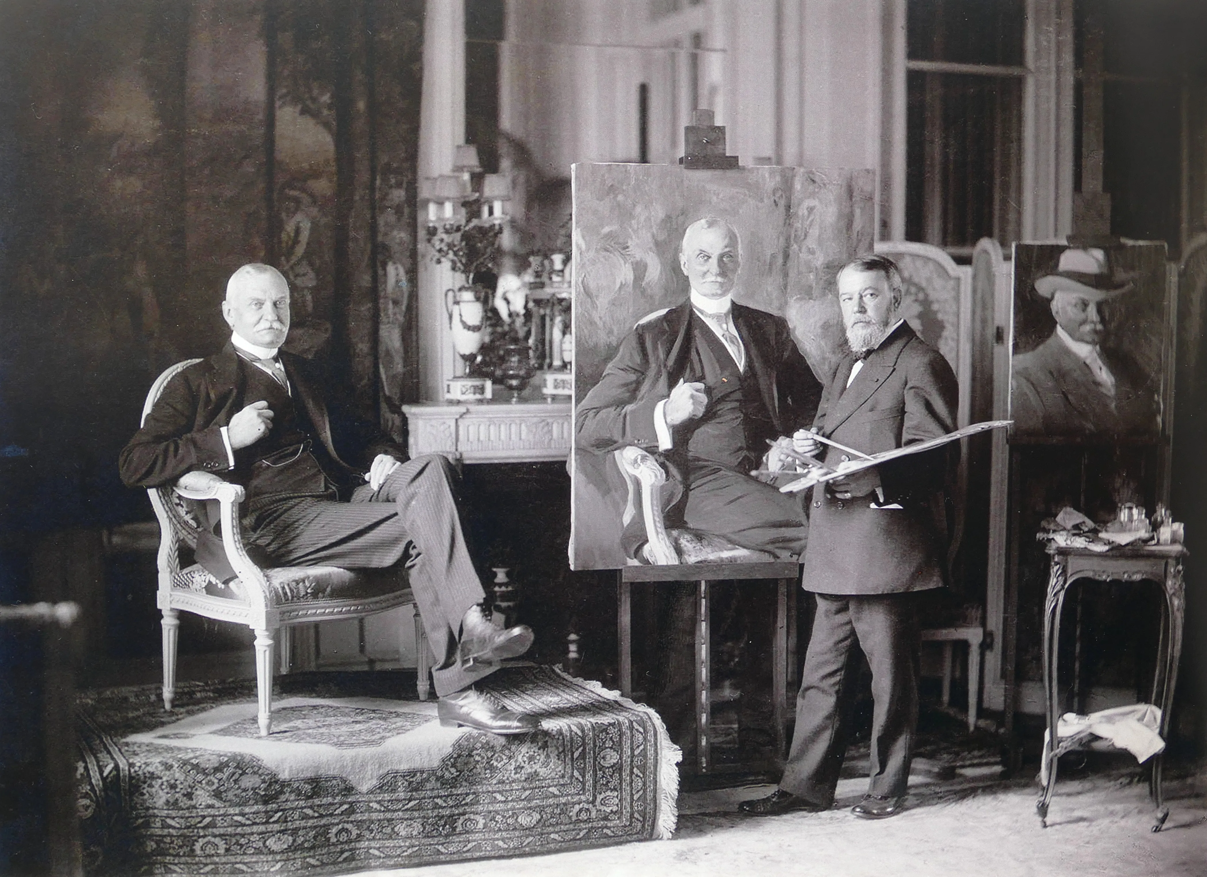 Twee mannen in een studio, de ene zittend en de andere staand met een penseel, omringd door kunstwerken en sierlijk meubilair