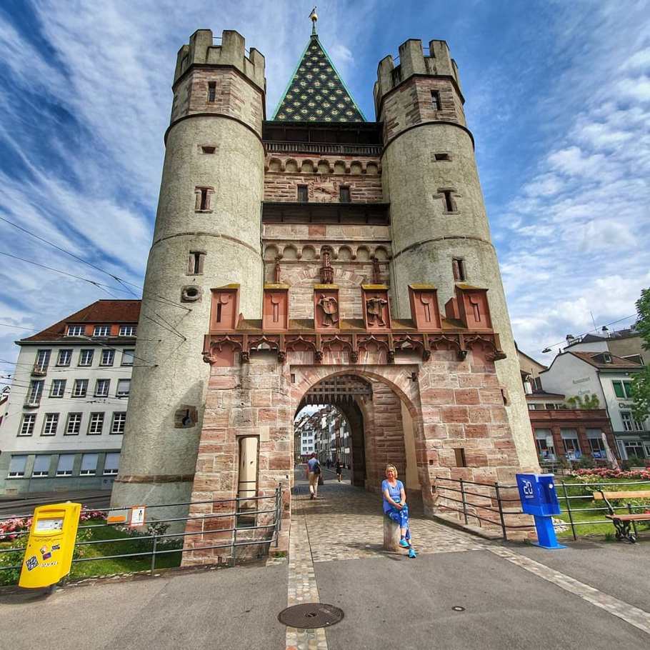 La Spalentor, una puerta medieval en Basilea, que cuenta con dos torres laterales redondas y una torre central cuadrada con techo verde, con gente caminando a través del arco.