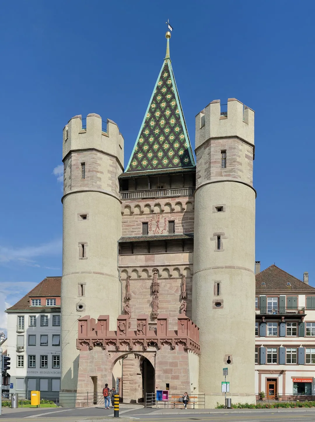 La puerta medieval Spalentor en Basilea, que presenta dos torres circulares a los lados, una torre central con un techo cónico de patrón verde y figuras esculpidas en la fachada de arenisca roja bajo un cielo azul despejado.