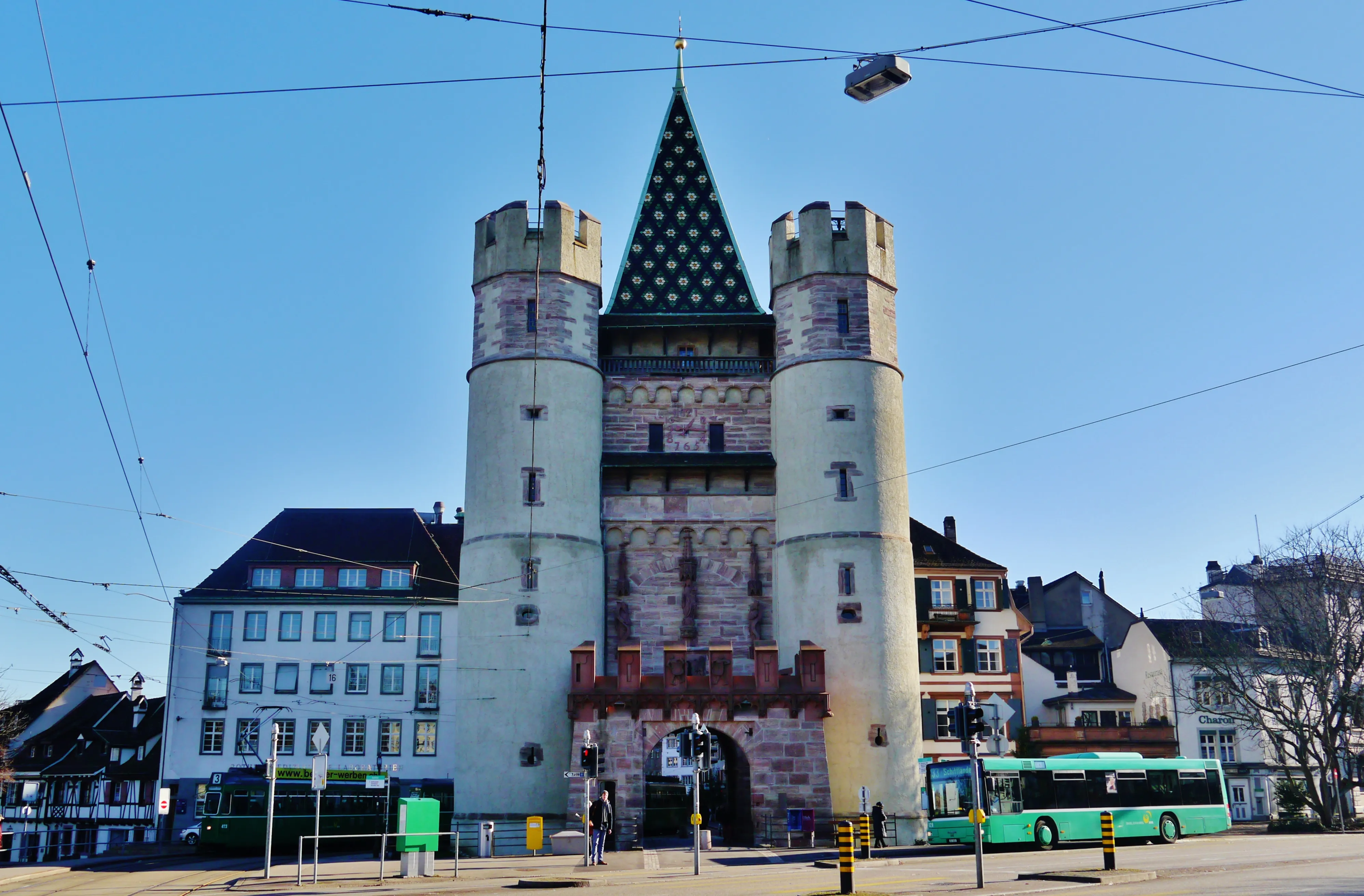 La puerta medieval Spalentor en Basilea, con una torre cuadrada central rematada por una aguja de azulejos verdes, dos torres laterales redondas y un arco de ladrillo rojo, sobre un cielo azul despejado con líneas de tranvía por encima y un autobús verde pasando.