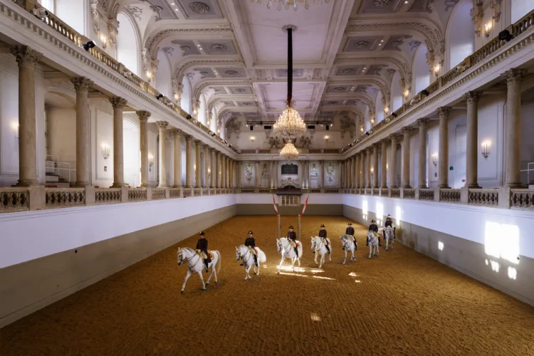 Riders on white horses in a grand, ornate riding hall with columns, balconies, and a chandelier.