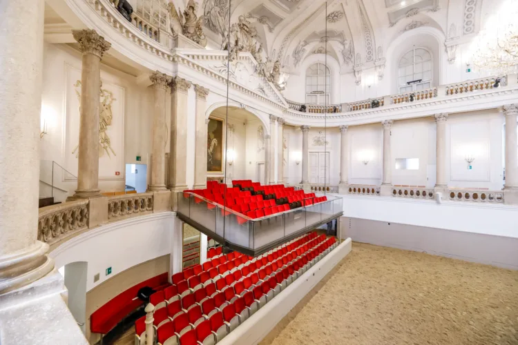 Interior ornamentado de un gran salón con asientos rojos, columnas blancas y un techo alto con molduras decorativas