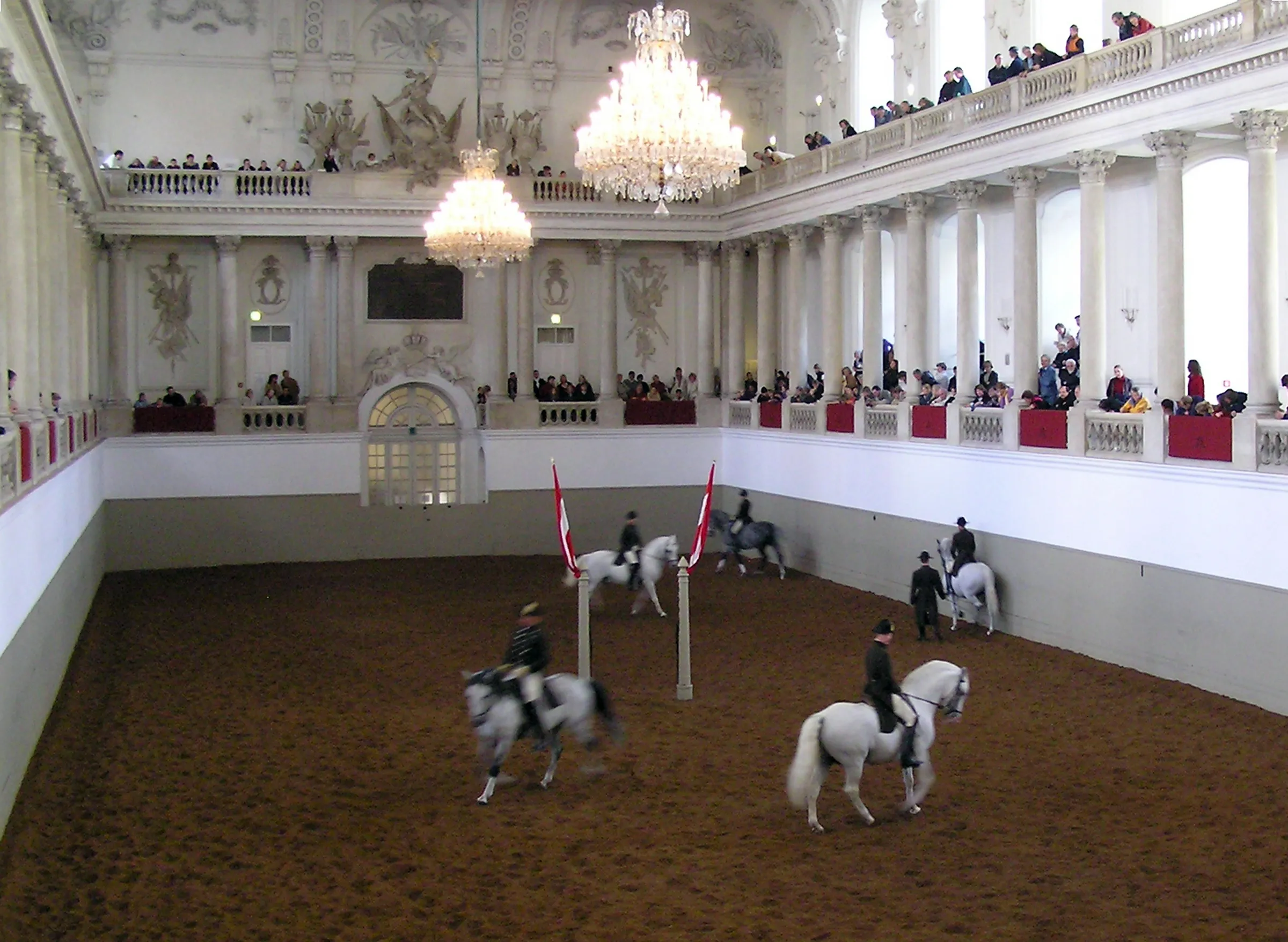 Riders on white horses perform in an ornate indoor arena with chandeliers and spectators on balconies