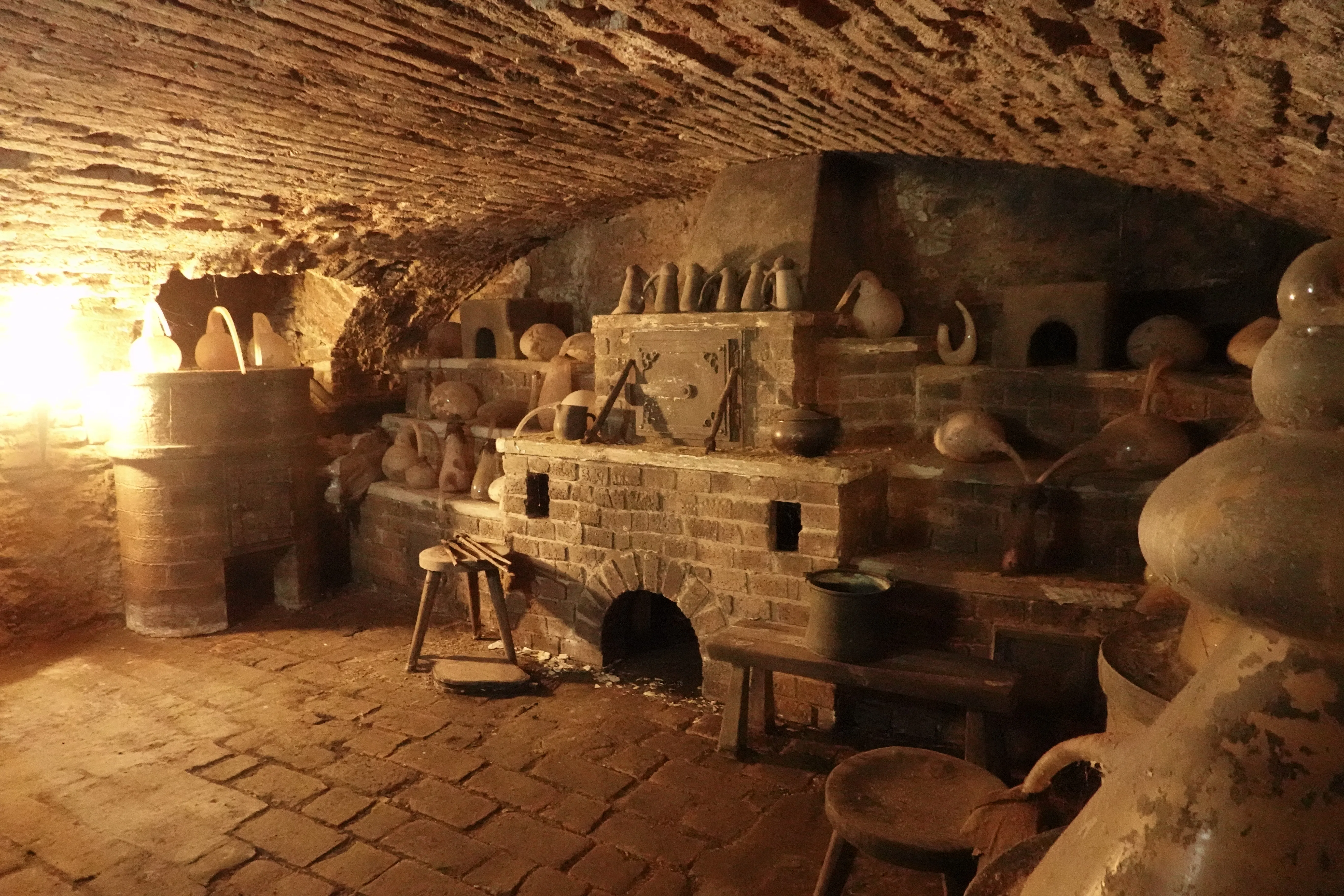 Ondergrondse kamer met stenen muren en een gewelfd plafond, met een nagebouwd alchemistisch laboratorium met vaten, een bakstenen oven en een werkbank.