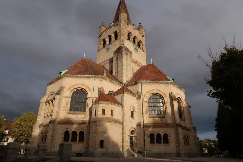Widok z zewnątrz na kościół św. Antoniego w Bazylei, ukazujący wysoką centralną wieżę ze spiczastą iglicą, dachy z czerwoną dachówką, duże łukowe okna i kamienną fasadę pod pochmurnym niebem