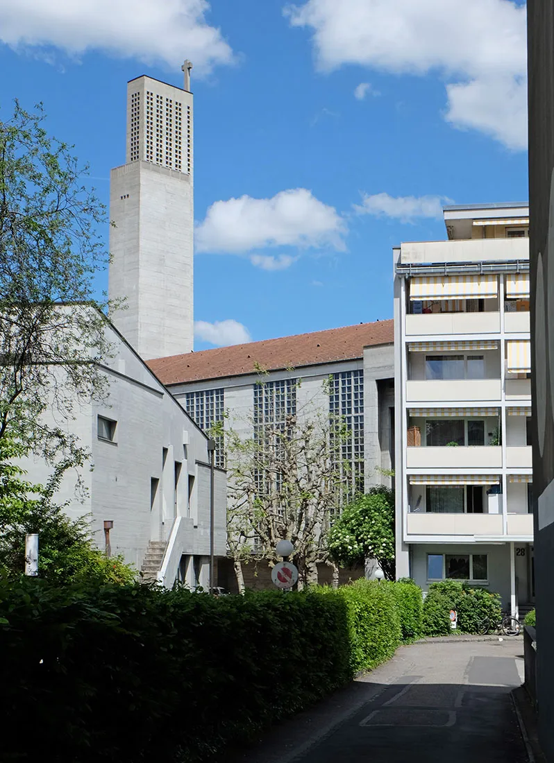 Wysoka betonowa dzwonnica z krzyżem wznosząca się nad kościołem St. Antonius w Bazylei, z dużymi oknami w kratę i przylegającym białym budynkiem mieszkalnym pod błękitnym niebem