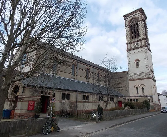 Widok z zewnątrz na kościół St Barnabas Church, obejmujący wysoką kamienną dzwonnicę z łukowatymi oknami, długą nawę z łukowatymi oknami, bezlistne drzewa oraz rower zaparkowany przy niskim płocie wzdłuż ulicy.