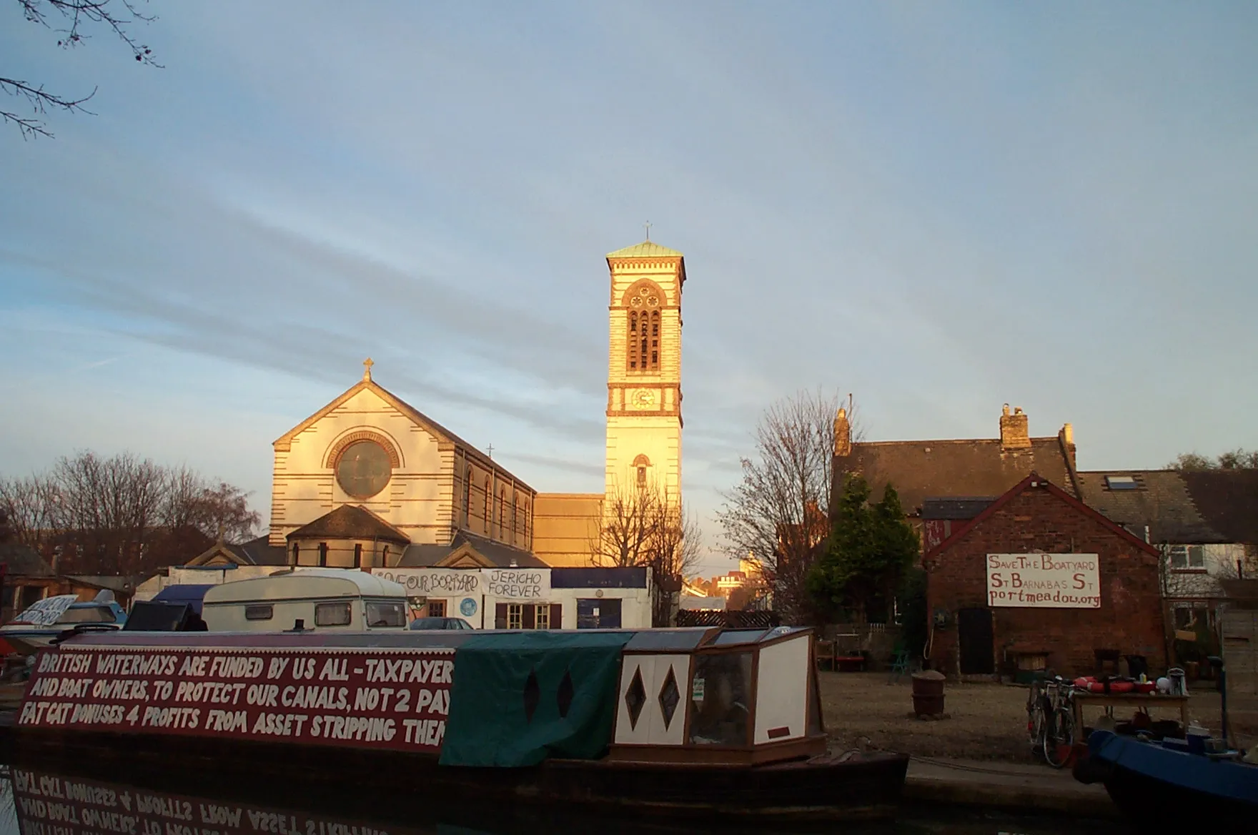 Kościół St Barnabas z wysoką dzwonnicą i okrągłym oknem rozetowym, widoczny wieczorem ponad kanałem z zacumowanymi na pierwszym planie łodziami typu narrowboat.