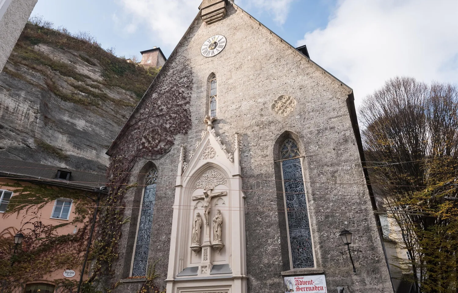 Stenen gotische kerkgevel met een klok bij de nok, hoge spitsboogramen met glas-in-lood en een centrale sculpturale crucifixnis, gelegen tegen een rotsachtige klif.