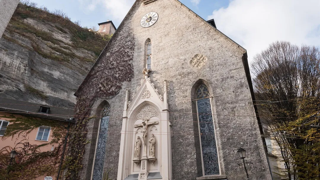 Stenen gotische kerkgevel met een klok nabij de topgevel, hoge smalle glas-in-loodramen en een gebeeldhouwde religieuze nis, gebouwd tegen een rotswand.