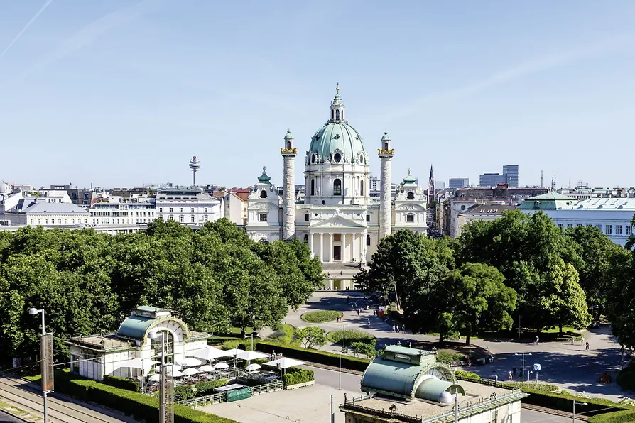 Iglesia de San Carlos con su cúpula verde y columnas gemelas, vista a distancia a través de Karlsplatz con árboles y edificios de la ciudad en primer y último plano
