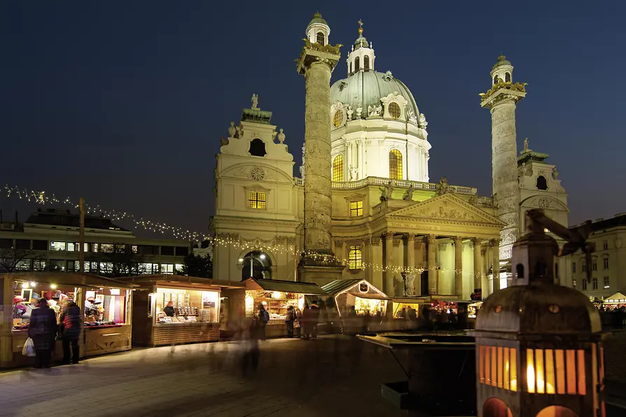 Iglesia de San Carlos por la noche con su cúpula verde y columnas gemelas iluminadas, puestos del mercado en primer plano