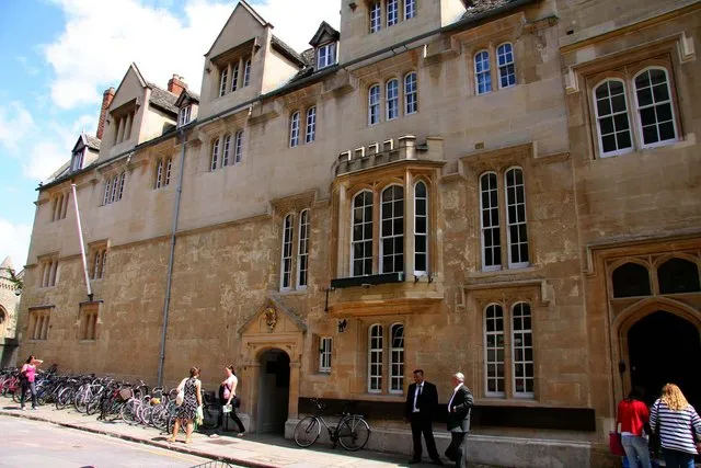 Fachada de piedra de St Edmund Hall con ventanas arqueadas, puerta de entrada y bicicletas aparcadas a lo largo de la calle con peatones pasando por delante