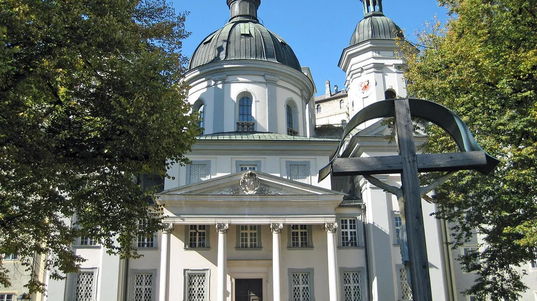 White Baroque church with a large dark dome and classical portico, framed by green trees, with a large dark metal cross sculpture in the foreground
