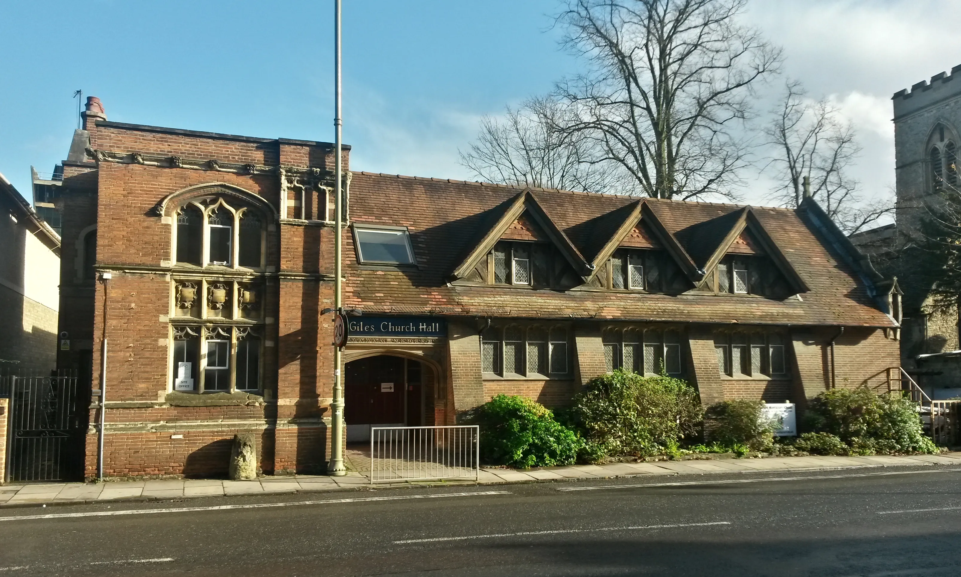 Buitenaanzicht van de roodbakstenen St Giles' Church Hall met een pannendak en boogramen, gelegen naast een stenen kerktoren.