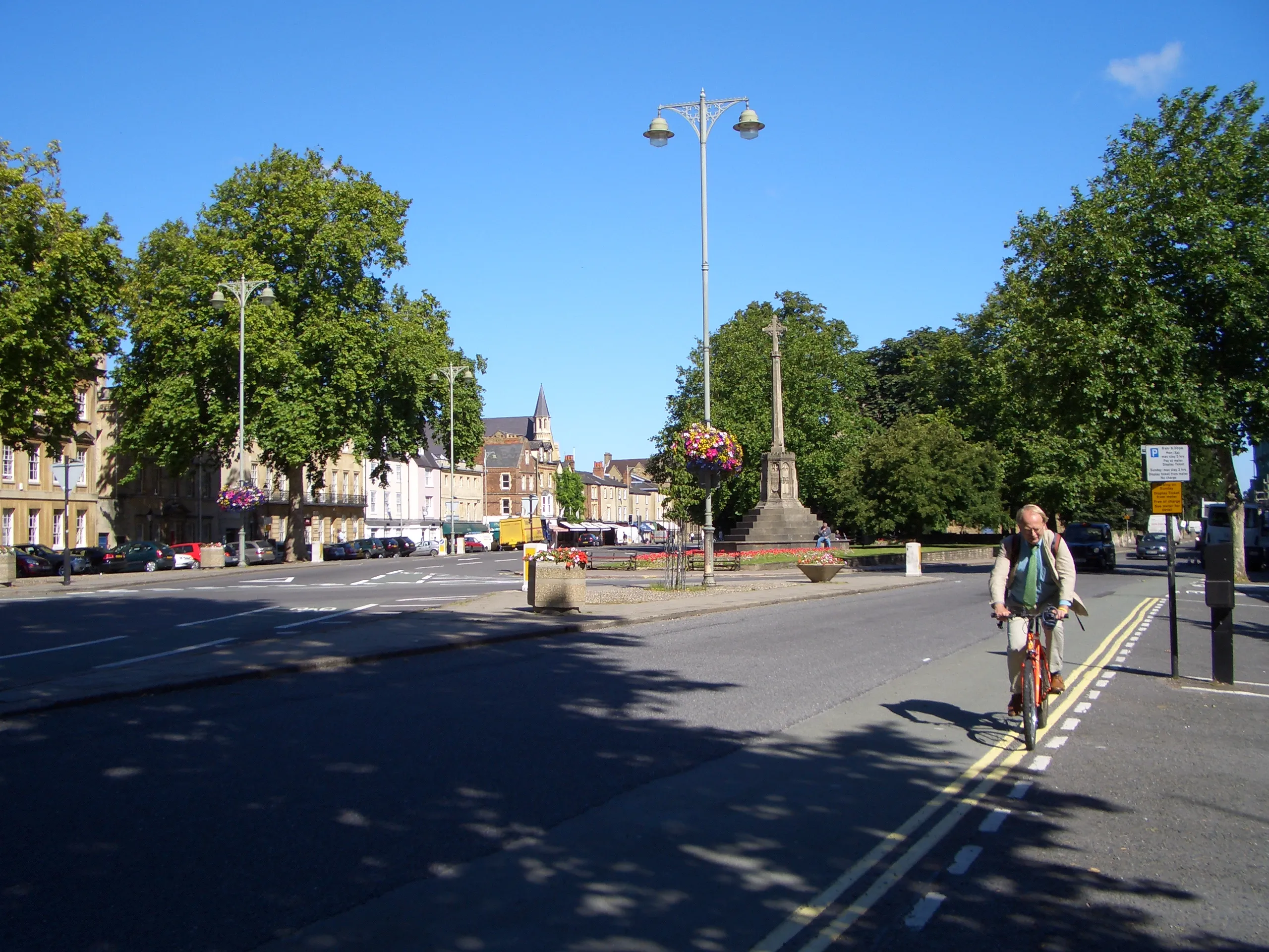 Straatbeeld in Oxford met een fietser op een weg met een middengeleider en een lantaarnpaal, waarbij de spits van St Giles' Church in de verte zichtbaar is onder een helderblauwe lucht.