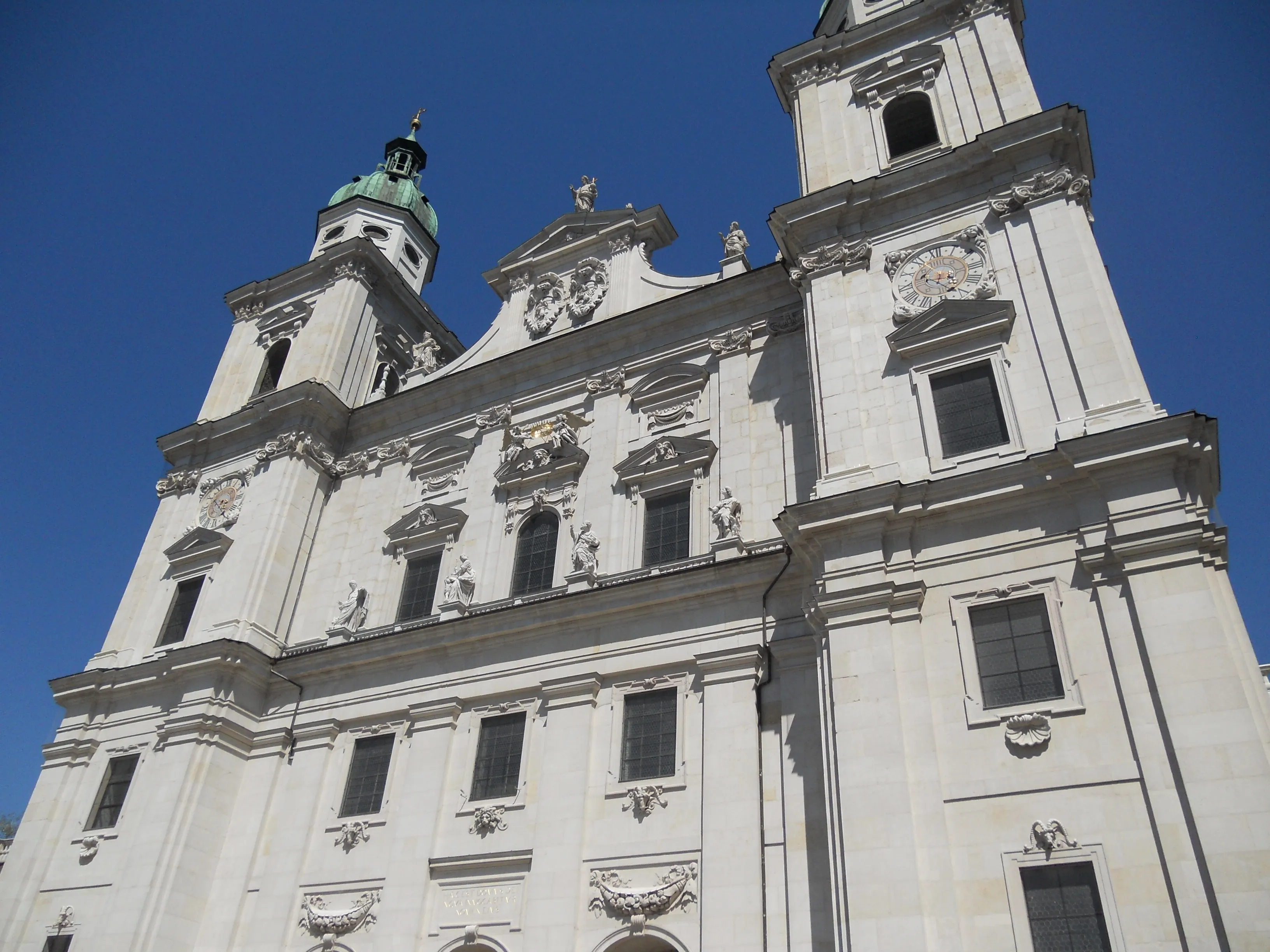 Biała barokowa fasada kościoła z dwiema wieżami i zieloną miedzianą kopułą na tle błękitnego nieba