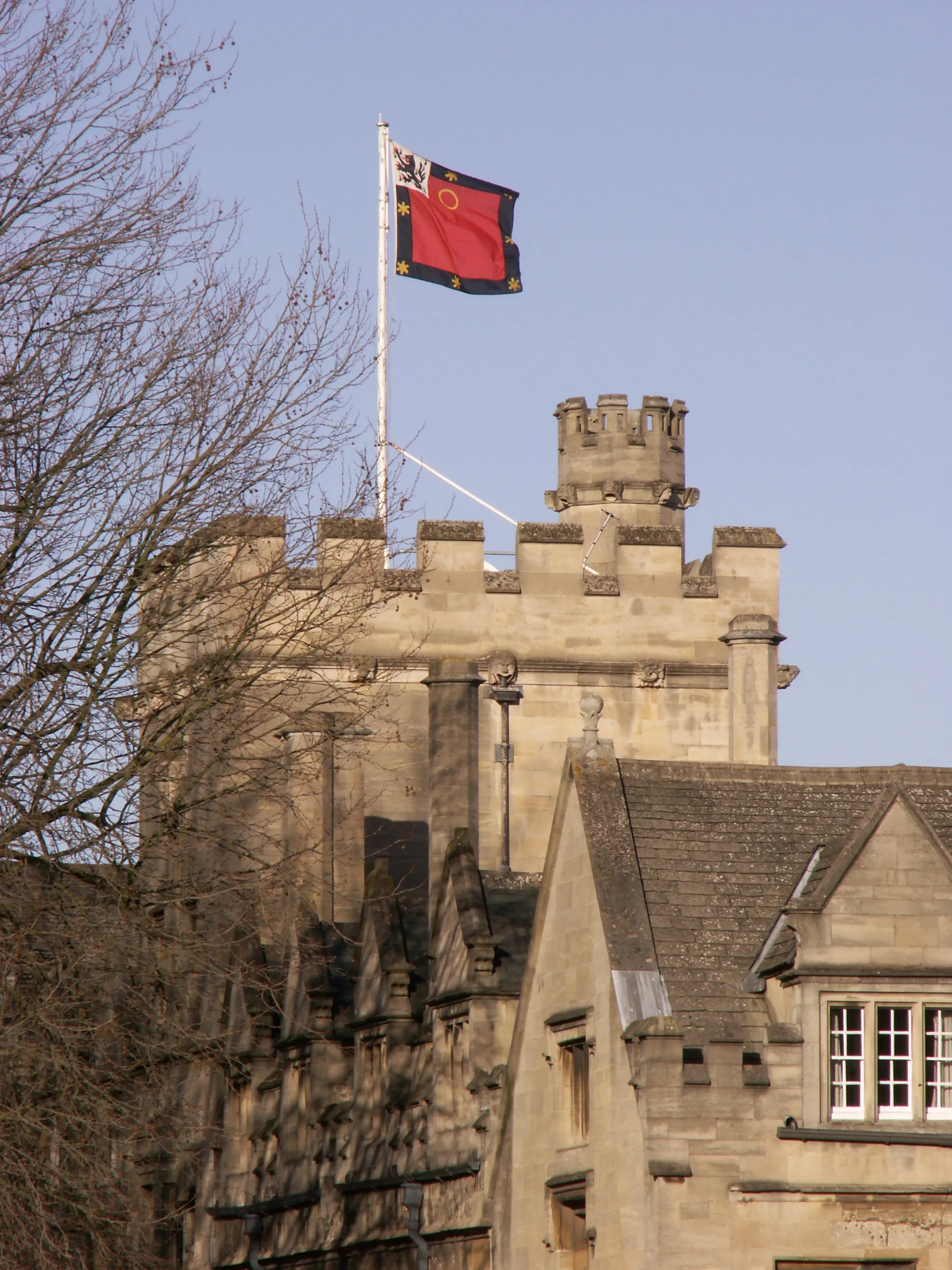 Stenen toren met gekanteelde borstweringen en een rood-zwarte heraldische vlag wapperend aan een vlaggenmast tegen een blauwe lucht, met kale takken op de voorgrond.