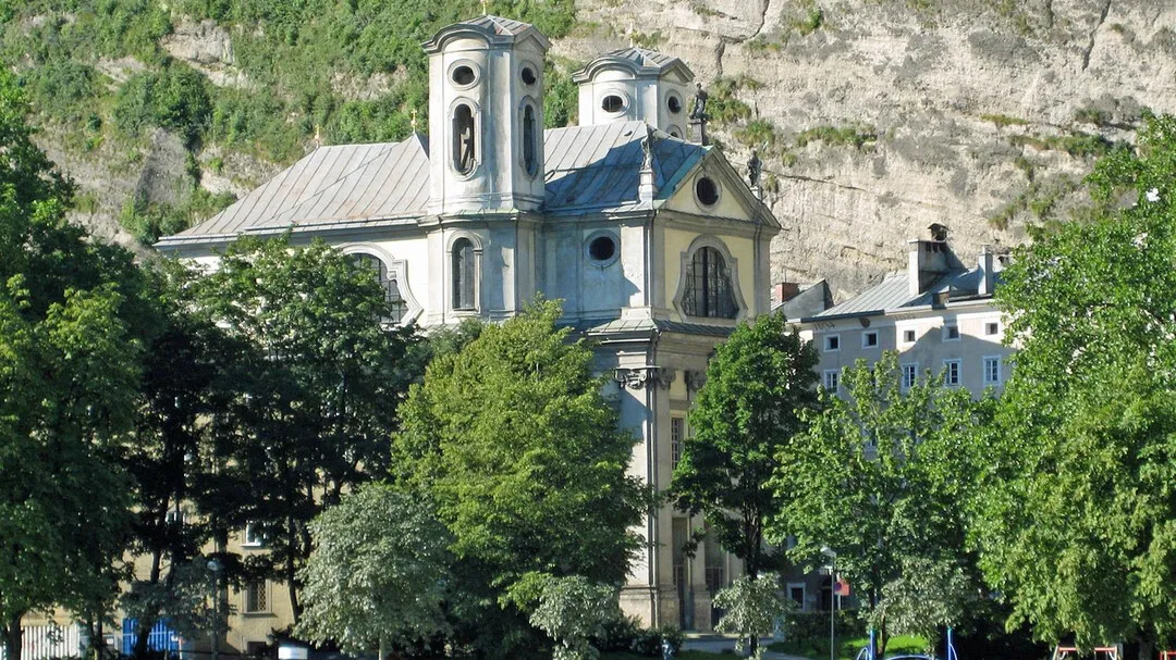 Exterior view of St. Mark's Church Salzburg with twin towers and a dome, set against a rocky cliff and surrounded by green trees.