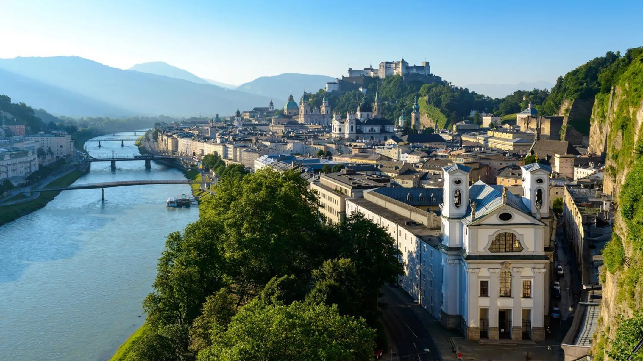 Aerial view of St. Mark's Church with its white facade and twin towers beside the Salzach River, with Hohensalzburg Fortress on the hill in the background.