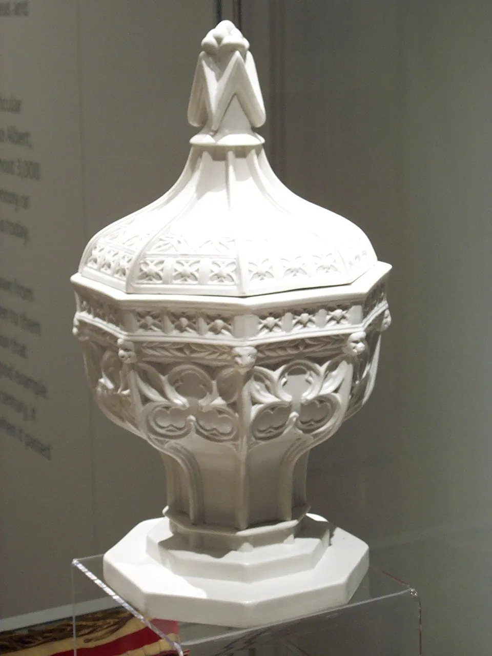 Pila bautismal de estilo gótico, de color blanco y ricamente tallada, con tapa decorativa y remate, expuesta sobre un soporte de acrílico transparente
