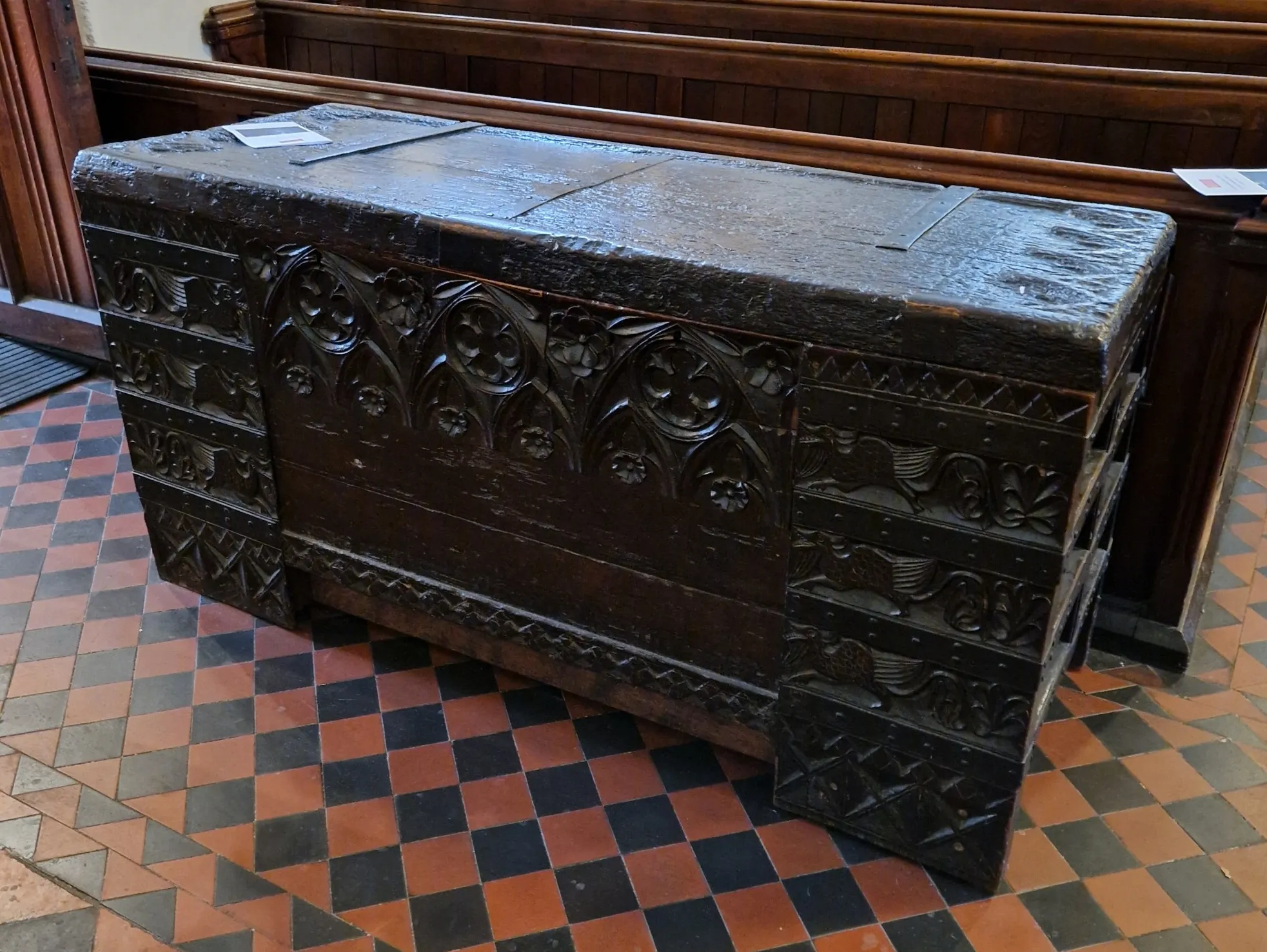 Un arcón de madera grande, oscuro y ornamentado, situado sobre un suelo a cuadros rojos y negros dentro de una iglesia, con bancos de madera al fondo.