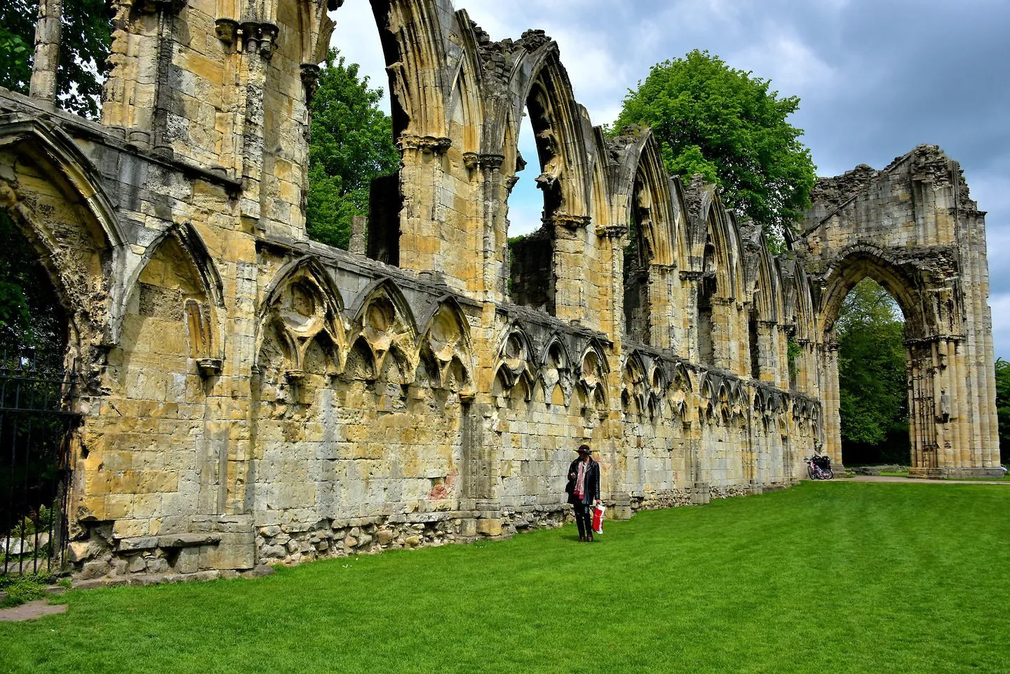 Vervallen stenen muur van St Mary's Abbey met een lange rij gotische bogen en arcades, met een persoon op een groen gazon ervoor voor de schaal, bomen en een bewolkte lucht op de achtergrond.