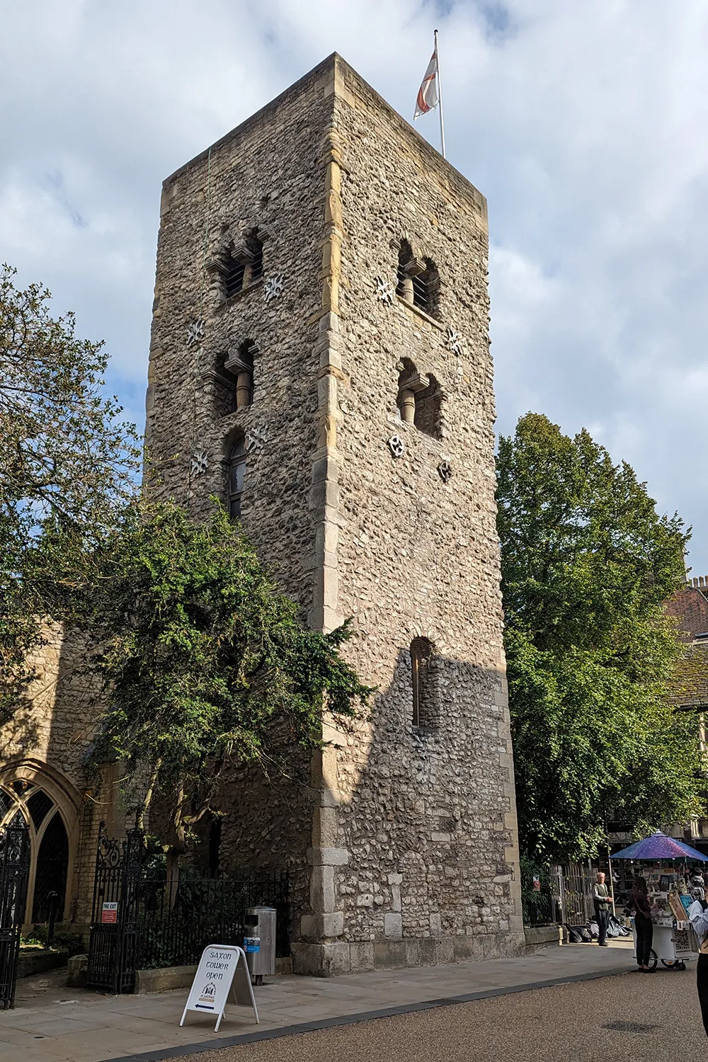 Hoge, rechthoekige Saksische stenen toren met ruw bewerkt metselwerk, boogvormige raamopeningen op meerdere niveaus, een vlag aan de top en klimop aan de linkerkant, met mensen en een marktkraam zichtbaar op straatniveau.