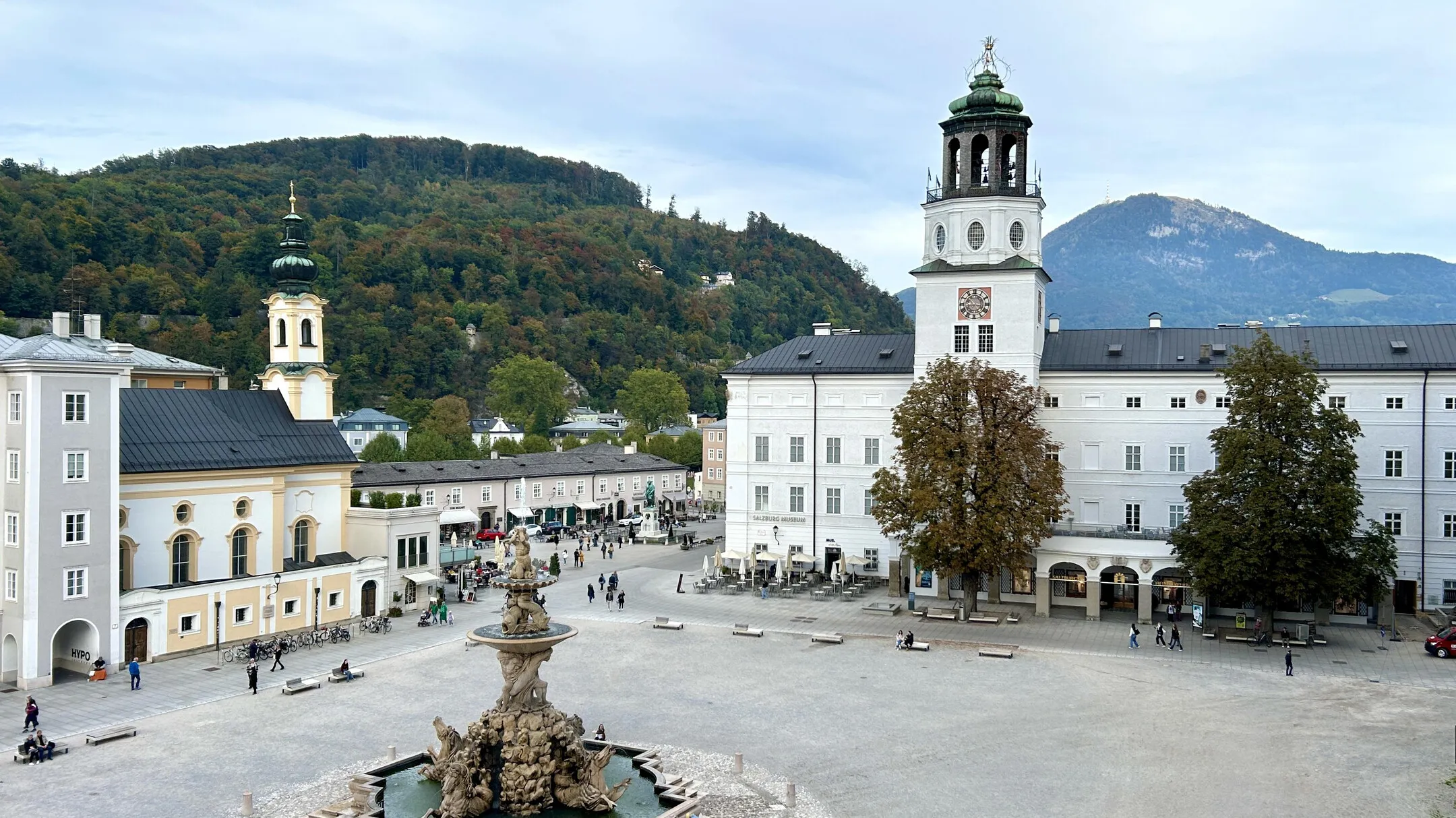 Vista panorámica de la Residenzplatz en Salzburgo mostrando la torre de la iglesia de San Miguel a la derecha, la ornamentada fuente Residenzbrunnen en primer plano, edificios barrocos y colinas boscosas al fondo