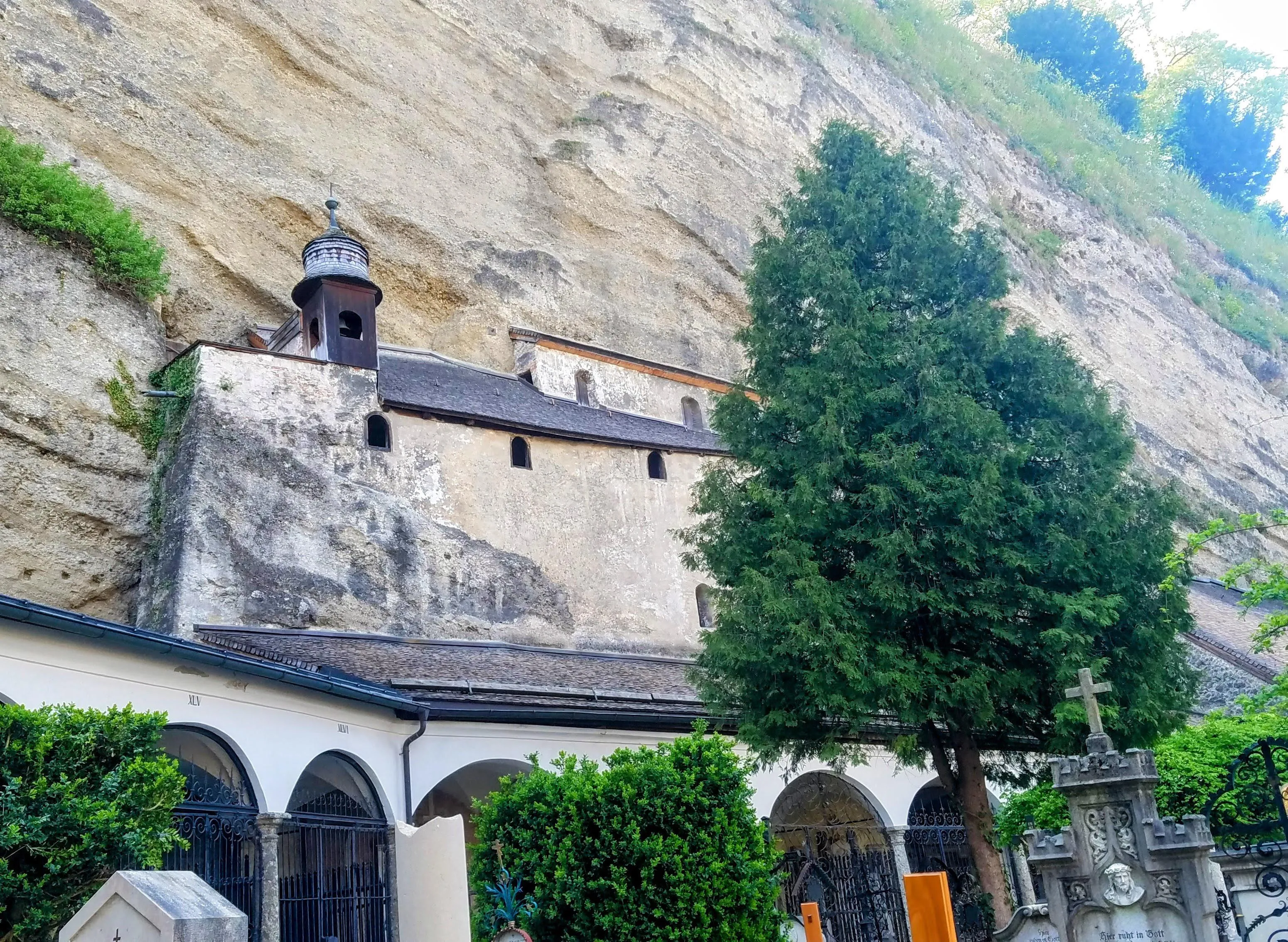 Una capilla con un pequeño campanario integrado en una pared rocosa, con columnatas arqueadas y un gran árbol en primer plano.