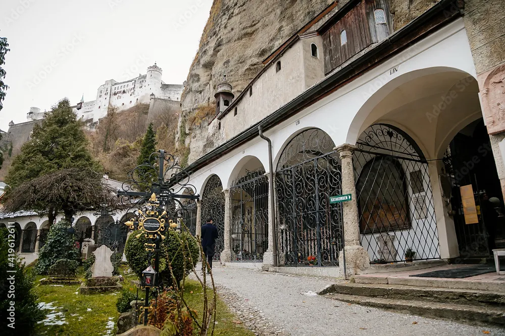 Arcada de piedra con ventanas de rejas de hierro construida en un acantilado, junto a un cementerio con lápidas, con una fortaleza blanca visible en la cima de la colina al fondo.