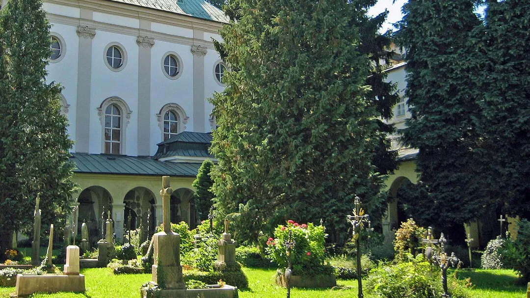 White baroque church facade with arched windows and arcaded cemetery containing gravestones, iron crosses, and tall green trees
