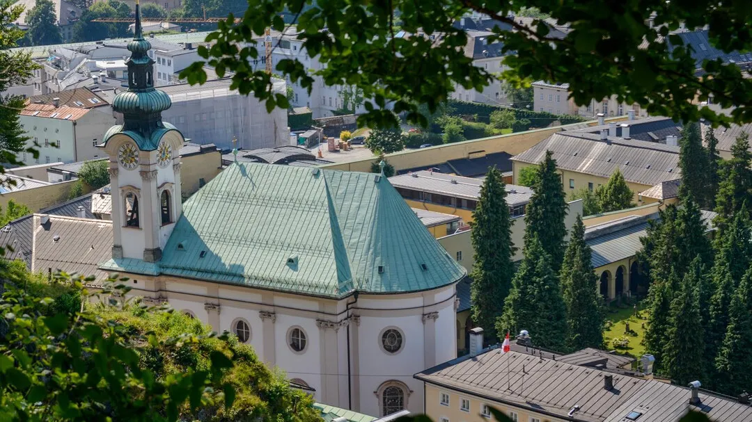 Aerial view of a church with a green copper roof and bell tower, surrounded by trees and an arcaded cemetery.