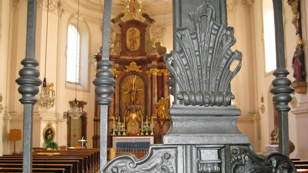 Interior of St. Sebastian's Church showing wooden pews, an ornate golden altar in the background, and a large decorative dark metal structure in the foreground.