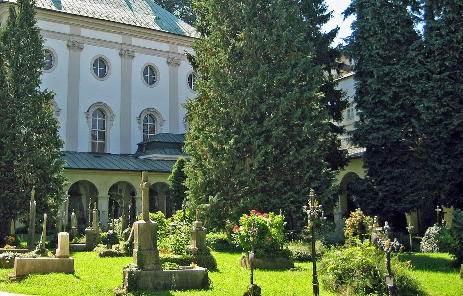 White church building with arched windows and a ground-floor arcade, viewed from a cemetery with gravestones on a green lawn surrounded by tall evergreen trees.
