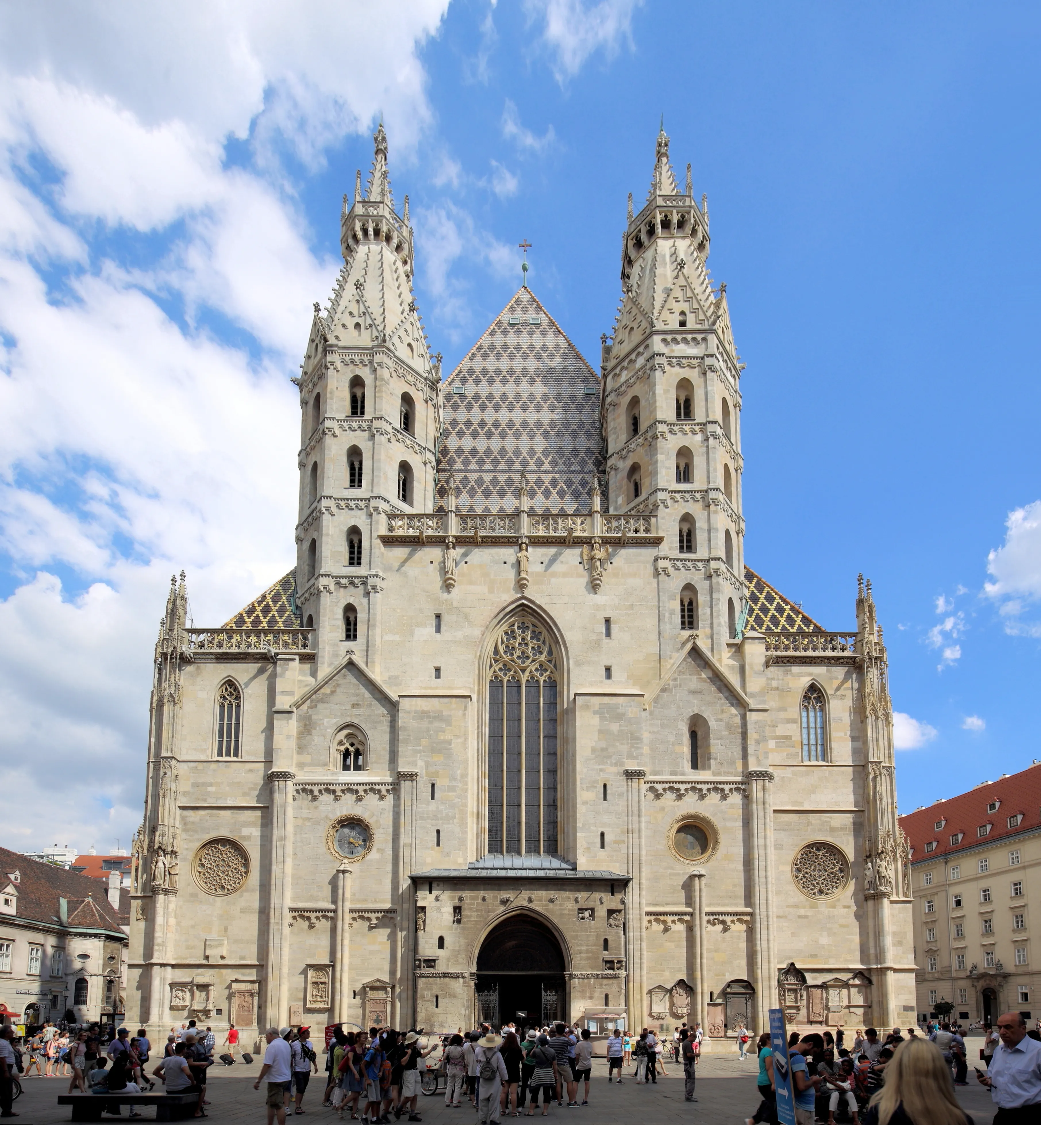 Front facade of St. Stephen's Cathedral in Vienna showing its twin Gothic towers, patterned tiled roof, and large central entrance with tourists gathered in the plaza below under a blue sky.