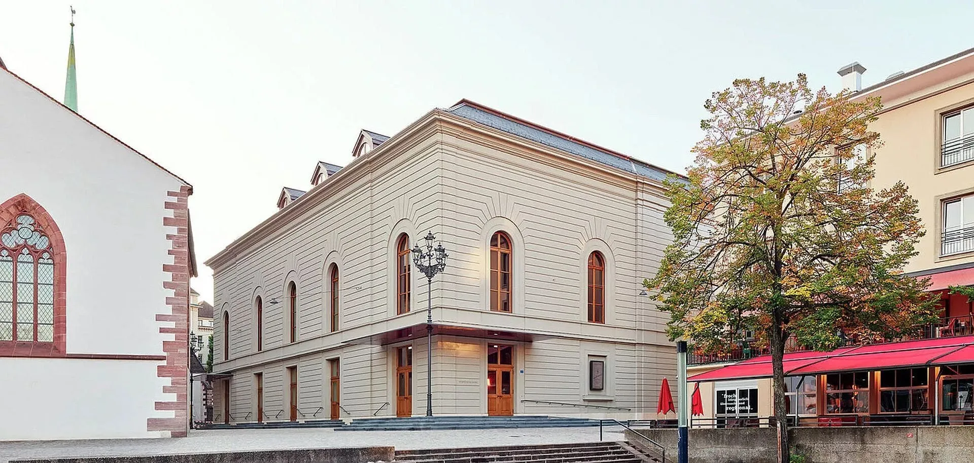 Exterior view of Stadtcasino Basel showing a light-colored classical building with arched windows, flanked by a church with a green spire on the left and a restaurant with red awnings on the right