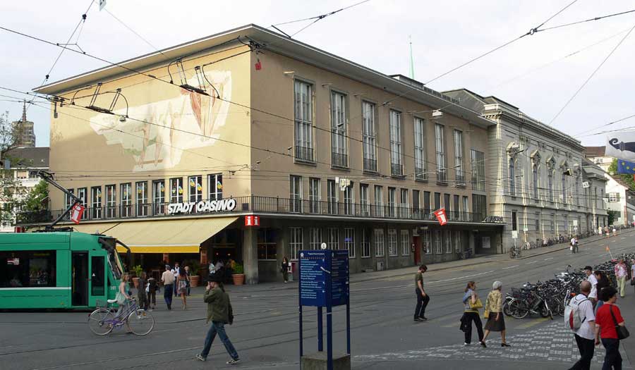 Exterior view of the Stadtcasino Basel building with a green tram passing in front and pedestrians walking on the plaza.