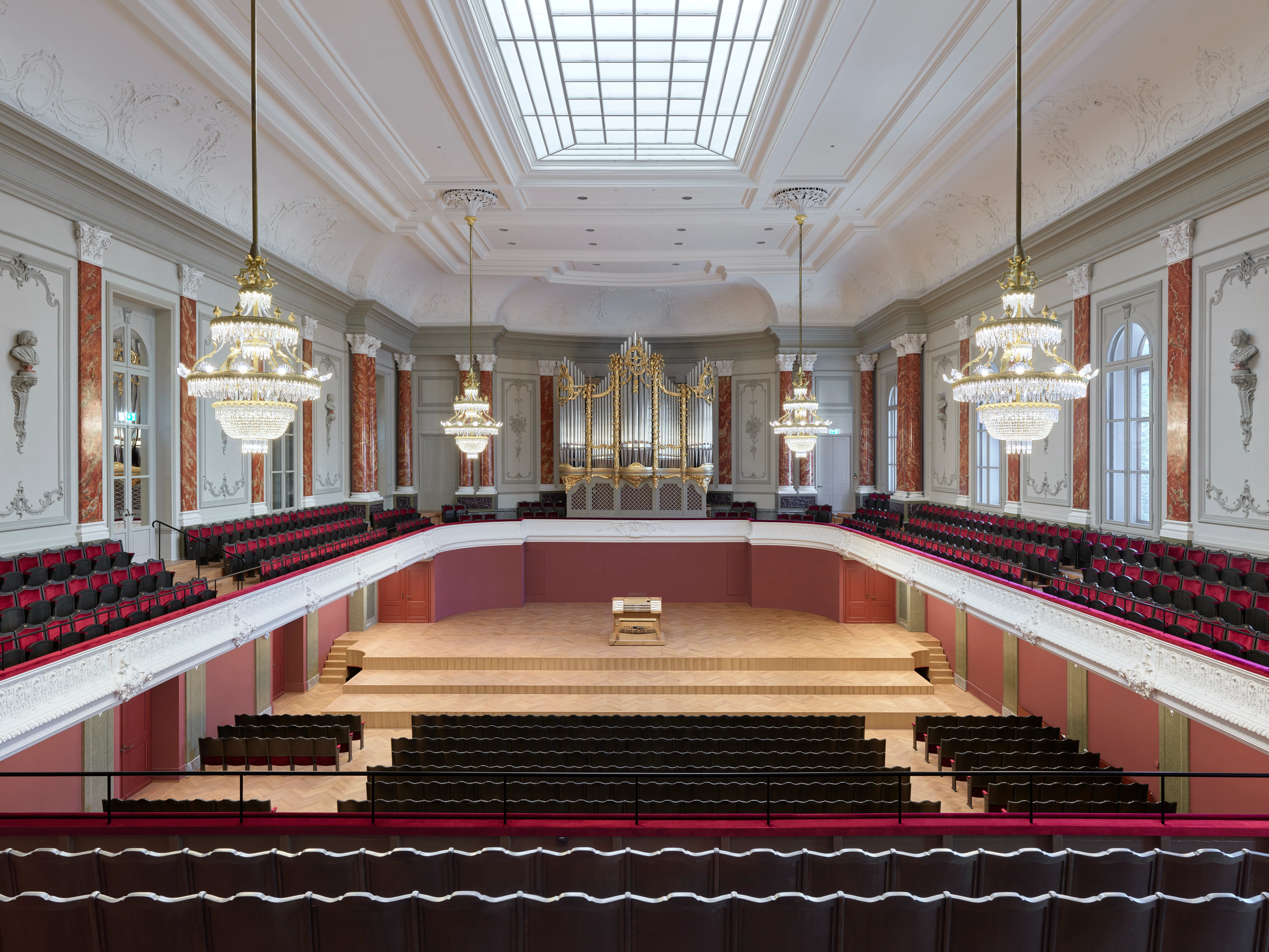 Wide interior view of the Musiksaal concert hall with rows of red velvet seating, hanging crystal chandeliers, a wooden stage, and a large pipe organ at the back wall beneath a skylight ceiling