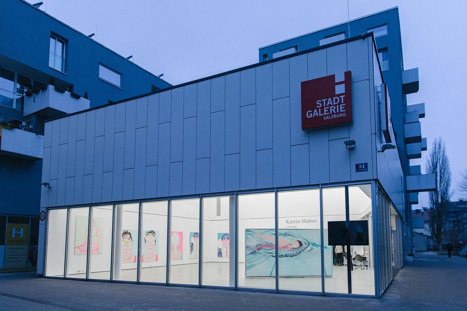Vista exterior del edificio de la Stadtgalerie am Lehen al atardecer, con un cartel cuadrado rojo con letras blancas, fachada de paneles blancos y grandes ventanales de cristal que muestran obras de arte en la planta baja.