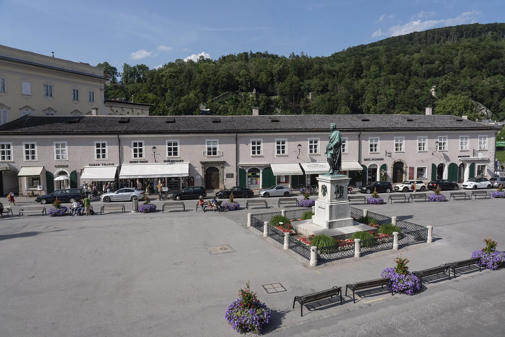 Wijd uitzicht op het Mozartplatz-plein in Salzburg met de lange witte gevel van het Stadtgalerie-gebouw, een centraal bronzen standbeeld op een voetstuk, bloembakken en een beboste heuvel op de achtergrond.