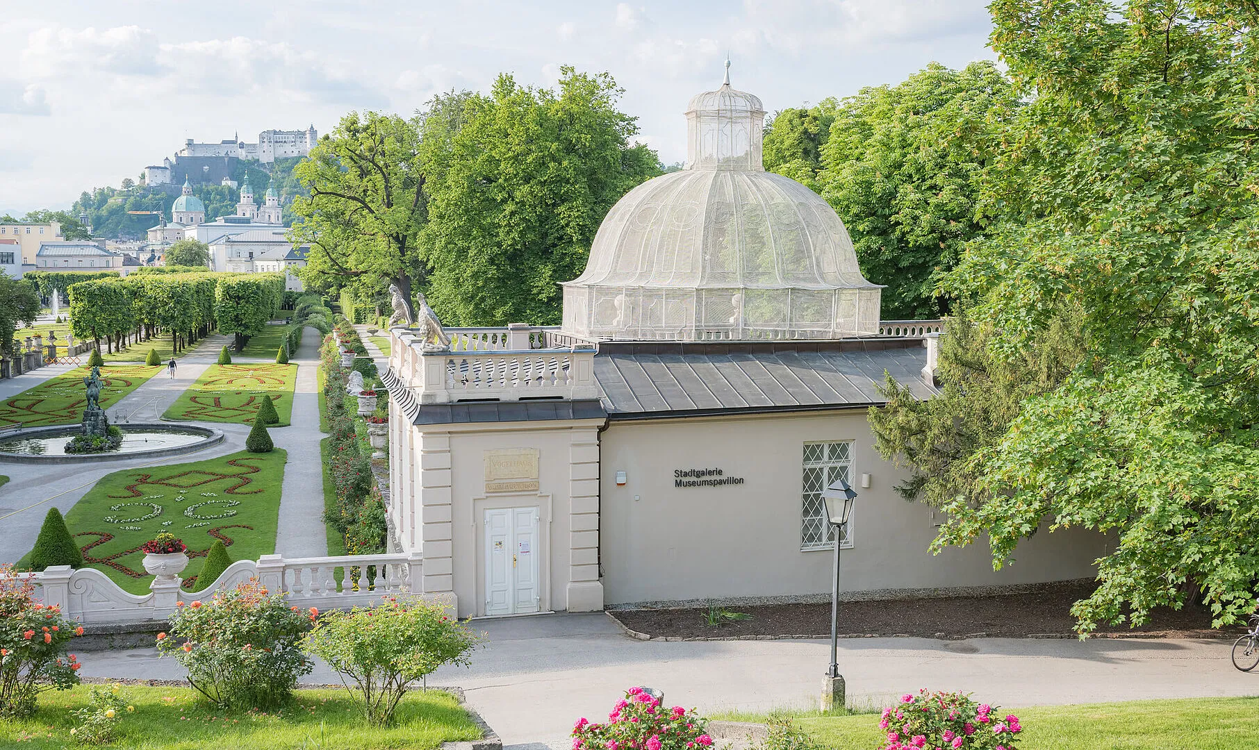 White domed pavilion building labeled Stadtgalerie Museumspavillon in a manicured garden with the Hohensalzburg Fortress on a hill in the background.