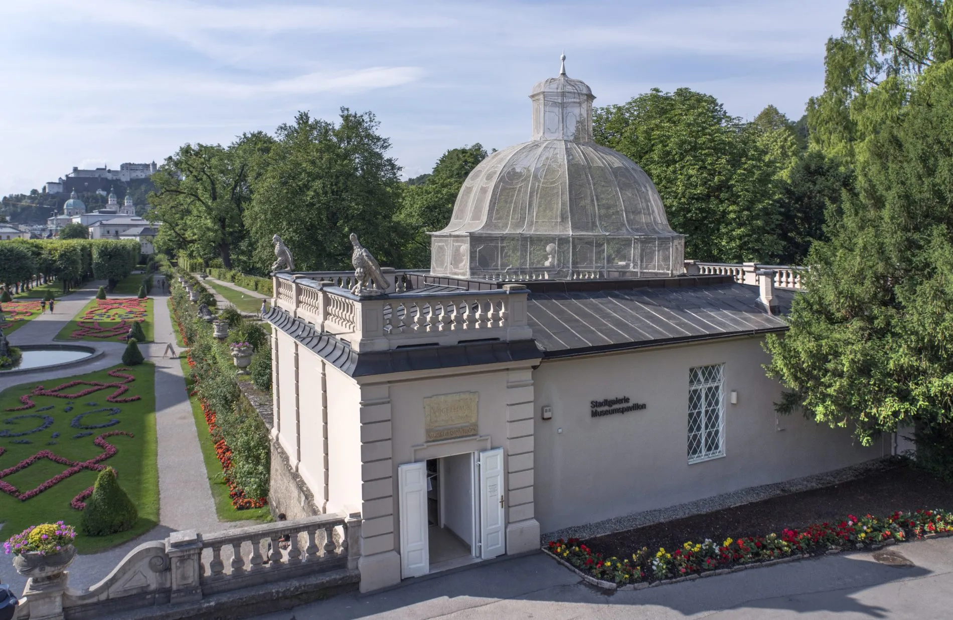 Exterior view of the Stadtgalerie Museumspavillon, a domed pavilion building with balustrade and statues, set within the Mirabell Gardens with formal flower beds and hedges