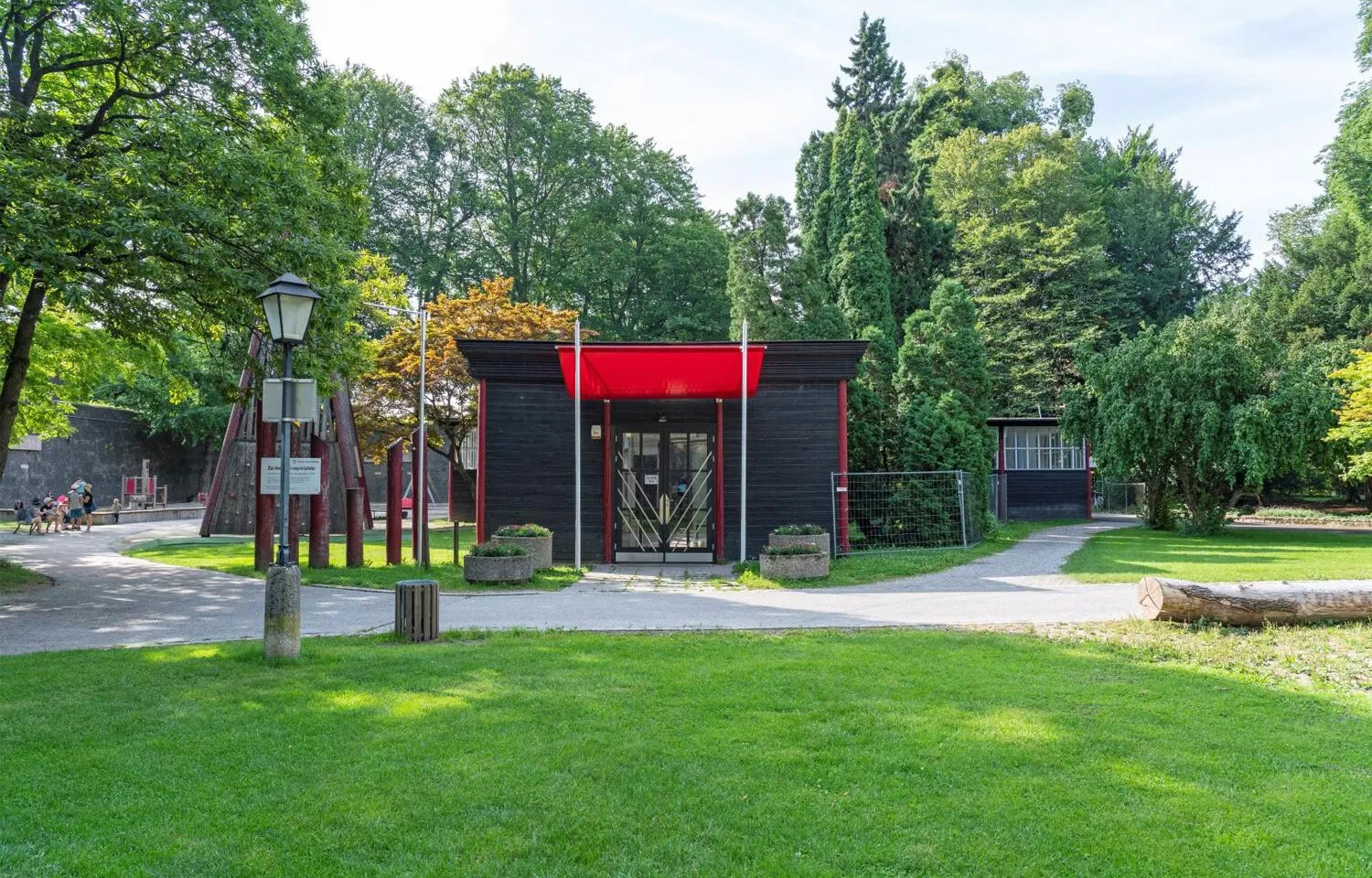 Un pequeño pabellón de madera oscura con un toldo rojo brillante sobre la entrada, situado en un parque verde con árboles, un césped y una farola antigua en primer plano.