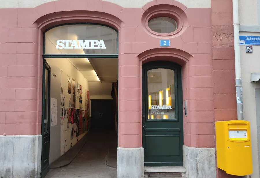 Różowa fasada STAMPA Galerie z łukowym wejściem, na którym widnieje nazwa galerii, zielonymi drzwiami z owalnym oknem oraz żółtą skrzynką pocztową po prawej stronie