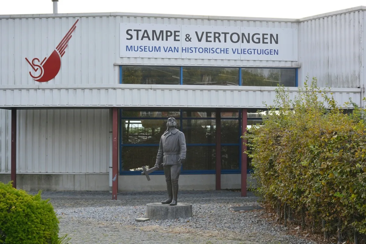 Widok z zewnątrz na Stampe & Vertongen Museum van Historische Vliegtuigen z pomnikiem lotnika z brązu przed wejściem, znakiem muzeum i czerwonym logo ze skrzydłami na fasadzie z blachy falistej w otoczeniu zielonych krzewów.
