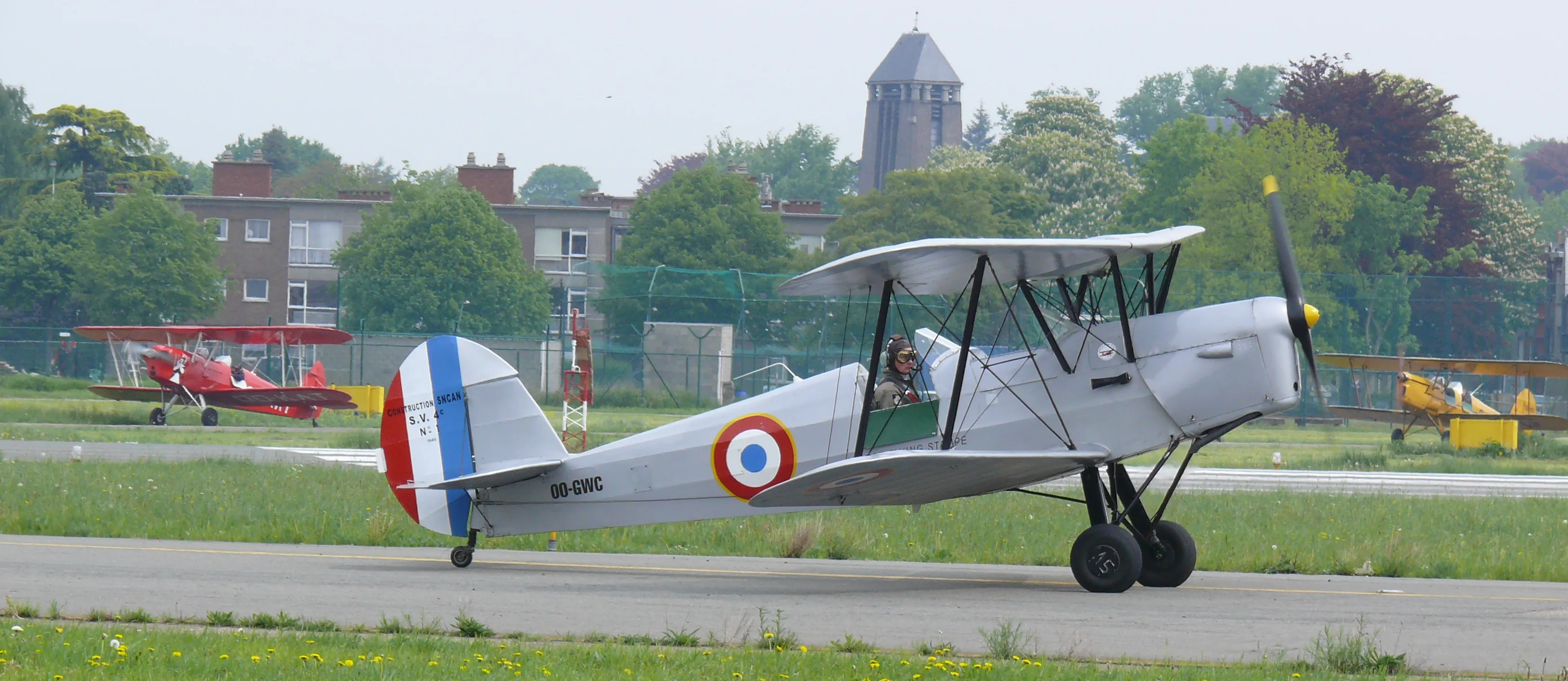Biplano Silver Stampe & Vertongen SV.4A con la escarapela belga y la cola con franjas rojas, blancas y azules carreteando por la pista, con aviones antiguos y una torre de agua al fondo