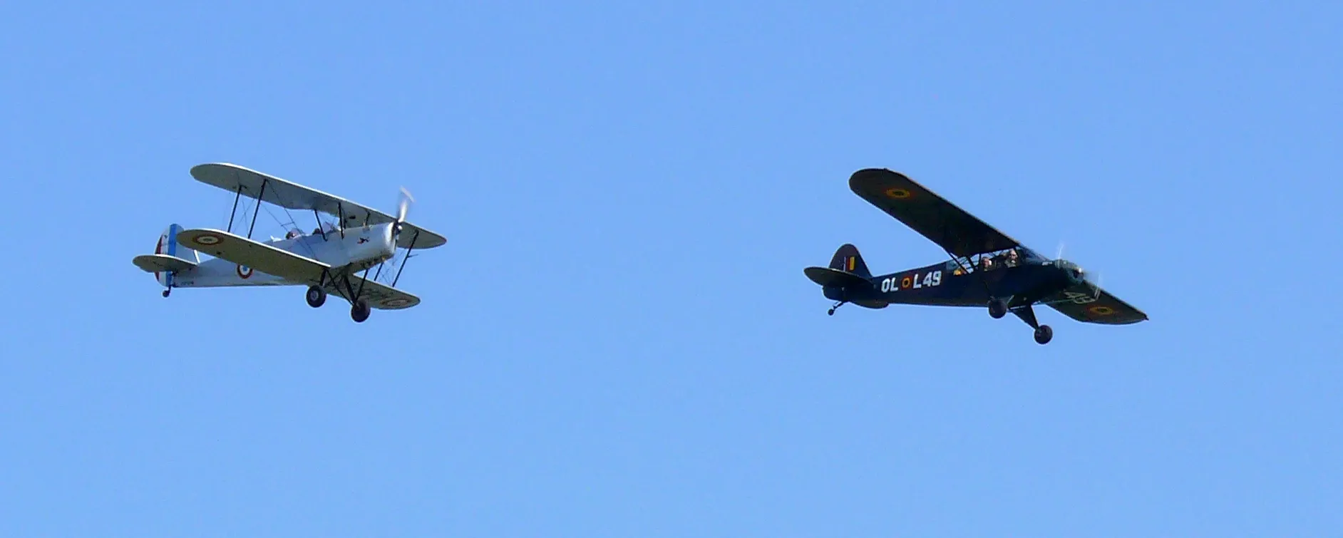 Dos aviones antiguos volando en formación contra un cielo azul despejado, con un biplano plateado a la izquierda y un monoplano oscuro a la derecha.